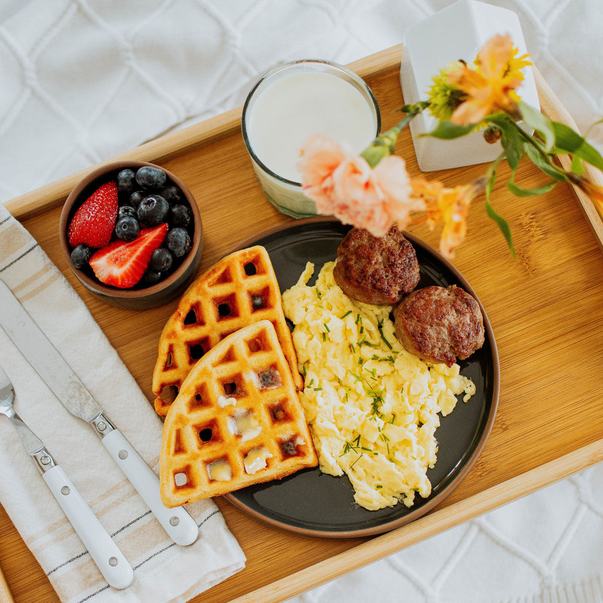 organic pork breakfast sausage on a plate with eggs and waffles on a tray with fruit, flowers, and milk